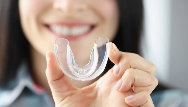 Smiling woman holding clear dental retainer or aligner in bright clinical setting.