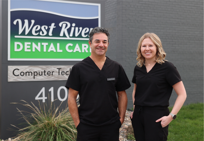 Two dental professionals in black scrubs stand outside West River Dental Care signage at their office entrance.